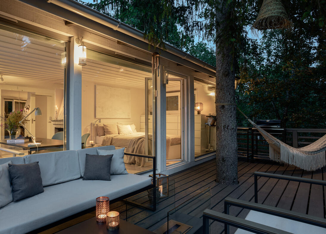 Woodland cabin terrace with outdoor seating, a hammock and warm interior light at twilight Woodland cabin terrace with outdoor seating, a hammock and warm interior light at twilight