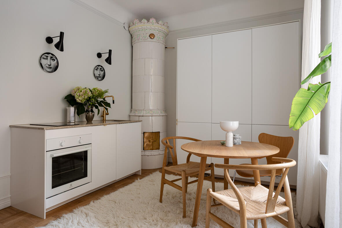 White kitchen and dining area with a tiled stove and sculptural Fornasetti wall plates. White kitchen and dining area with a tiled stove and sculptural Fornasetti wall plates.